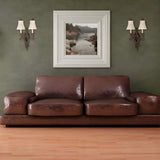 A spacious room features a large brown leather sofa set against a green wall, embodying traditional style. Above the sofa hangs a framed landscape painting of a serene lake and mountains, flanked by two Drawing-1 Room 2 Light Wall Lights with walnut finish and white lampshades. The light wood floor has a walnut finish.