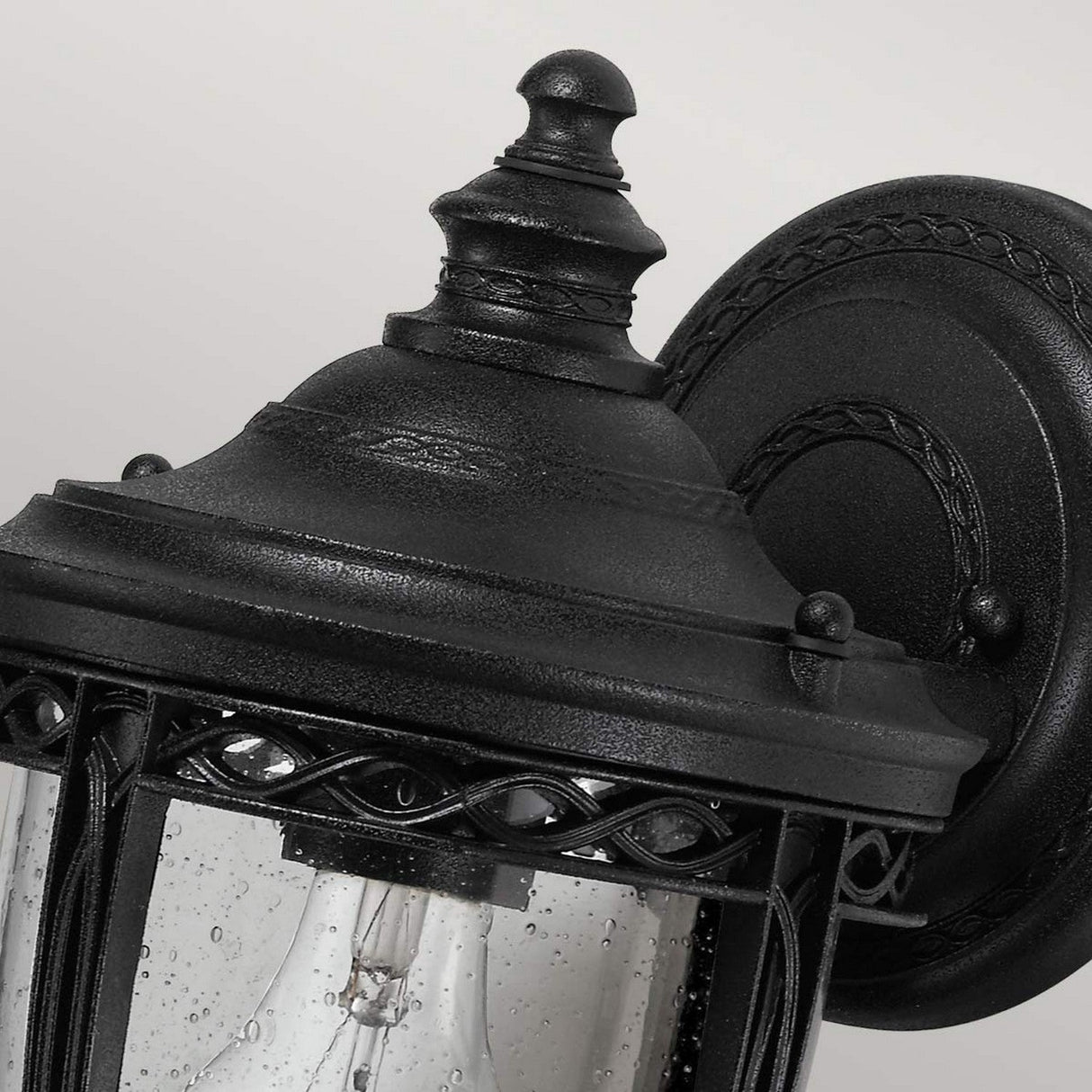 Close-up of the English Bridle 1 Light Outdoor Small Wall Lantern in a black finish, showcasing its vintage-inspired design with ornate detailing and a clear glass shade that reveals the lightbulb inside, set against a simple light gray background.