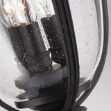 Close-up of the English Bridle 3 Light Outdoor Large Pedestal - Black with raindrops on its clear glass surface. The sleek black finish frame encloses lit bulbs, casting a cozy, warm glow against the droplets, capturing the essence of refined outdoor lighting in a rainy scene.