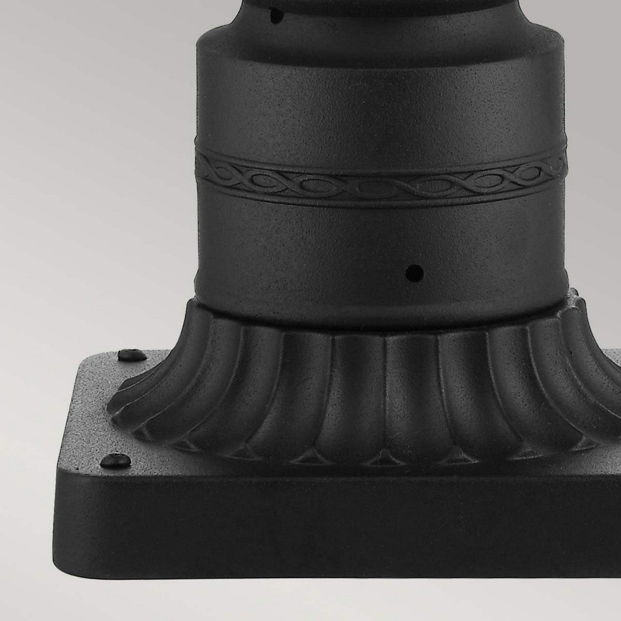 Detailed focus on a black metal base with intricate decorative elements, displaying a fluted design along with an engraved band. It is mounted on a square platform secured by four small bolts at each corner. This design pairs beautifully with the English Bridle 3 Light Outdoor Medium Pedestal in black finish to highlight its elegance.