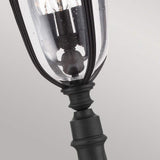 A detailed view of the English Bridle 3 Light Outdoor Medium Pillar - Black highlights its vintage-style outdoor charm with a black metal frame. The clear seeded glass cover displays multiple bulbs inside, set against a light gray backdrop.