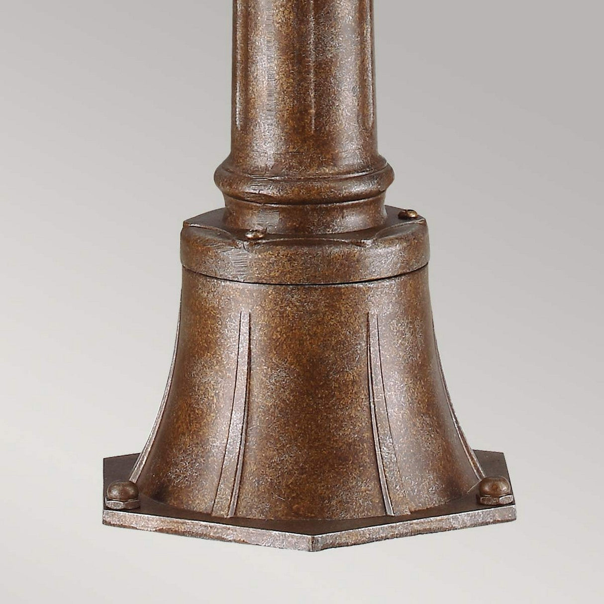 Here's a revised sentence incorporating the product data:

Close-up of the English Bridle 3 Light Outdoor Medium Pillar Lantern - Bronze, showcasing its bronze-colored metal base with a flared, textured design. The cylindrical base gracefully tapers upwards and is secured with small bolts, while clear seeded glass enhances the piece against a simple light gray background.