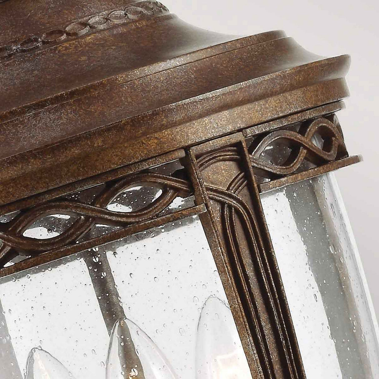 A close-up of the English Bridle 3 Light Outdoor Large Chain Lantern - Bronze showcases its vintage-style elegance, crafted with a British Bronze finish. This ornate design features glass panels enhanced with decorative accents, glistening with water droplets that suggest its resilience to the elements as outdoor lighting.