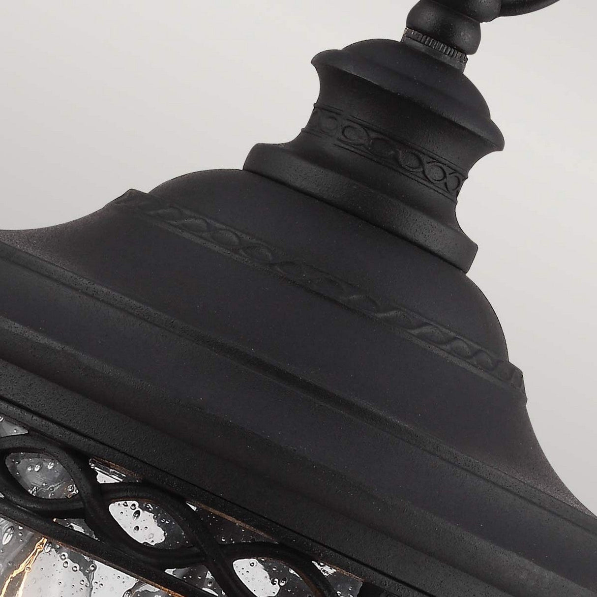 A detailed view of the English Bridle 3 Light Outdoor Medium Chain Lantern - Black showcases its vintage flair. This outdoor lighting fixture features a decorative top and a metal frame with intricate patterns, while its textured bubbled glass elevates its elegant design, adding timeless charm to any setting.