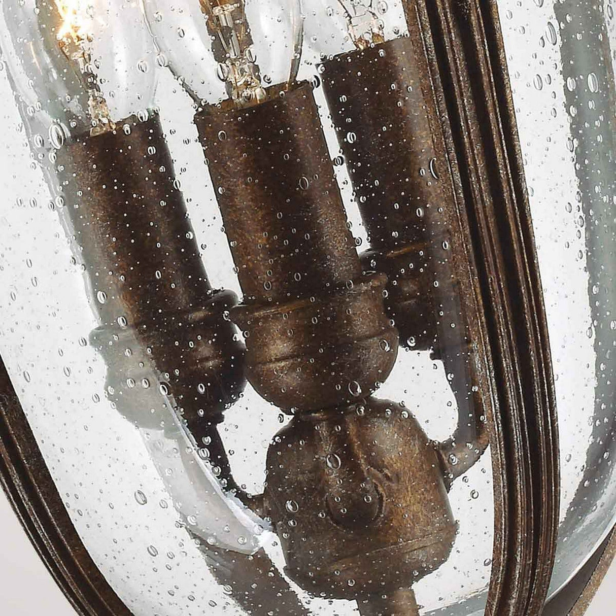 A close-up of the English Bridle 3 Light Outdoor Medium Chain Lantern showcases three vintage-style light bulbs enclosed within a seeded glass casing adorned with water droplets, achieving a rustic and elegant appearance. The fixture’s metal components gleam in a charming bronze finish.