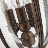 A close-up of the English Bridle 3 Light Outdoor Medium Chain Lantern showcases three vintage-style light bulbs enclosed within a seeded glass casing adorned with water droplets, achieving a rustic and elegant appearance. The fixture’s metal components gleam in a charming bronze finish.