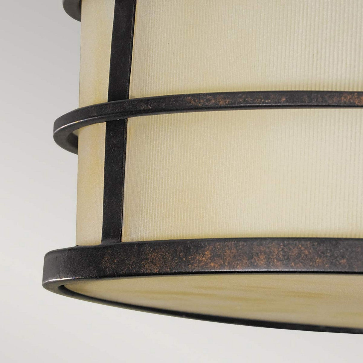 A close-up of the Fusion 3 Light Semi Flush in Grecian Bronze reveals its beige cylindrical pendant design with dark metal accents. Two concentric Grecian Bronze metal bands encircle the lamp, highlighting a rustic and industrial style against a light gray background, perfect for enthusiasts of semi flush lighting designs.