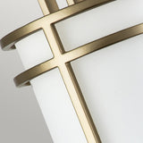 A close-up of the Fusion 1 Light Wall Light - Painted Natural Brass features a cylindrical white shade adorned with overlapping gold metal bands in a geometric design, with the painted natural brass providing an elegant contrast to the soft gray background.