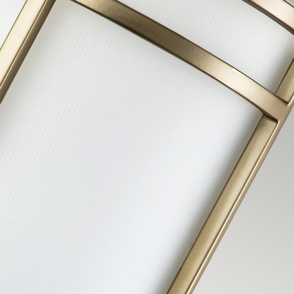 Close-up of a modern light fixture showcasing the Fusion 1 Light Wall Light in Painted Natural Brass with a frosted glass panel and brushed metal frame. Its curved design, along with metallic elements and a ribbed opal glass shade, delivers a sleek and elegant appearance.