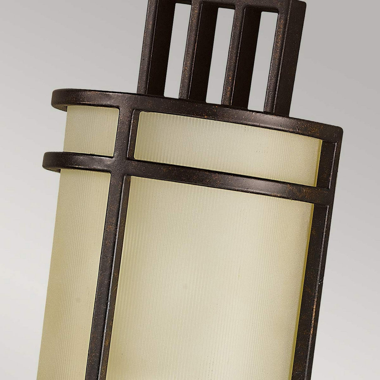 Close-up of the Fusion 1 Light Wall Light in Grecian Bronze, a modern cylindrical outdoor wall sconce. It features a frame with vertical and horizontal lines that encase a ribbed glass shade for soft light diffusion. The fixture showcases a contemporary design with clean, geometric elements and subtle Arts & Crafts influences.