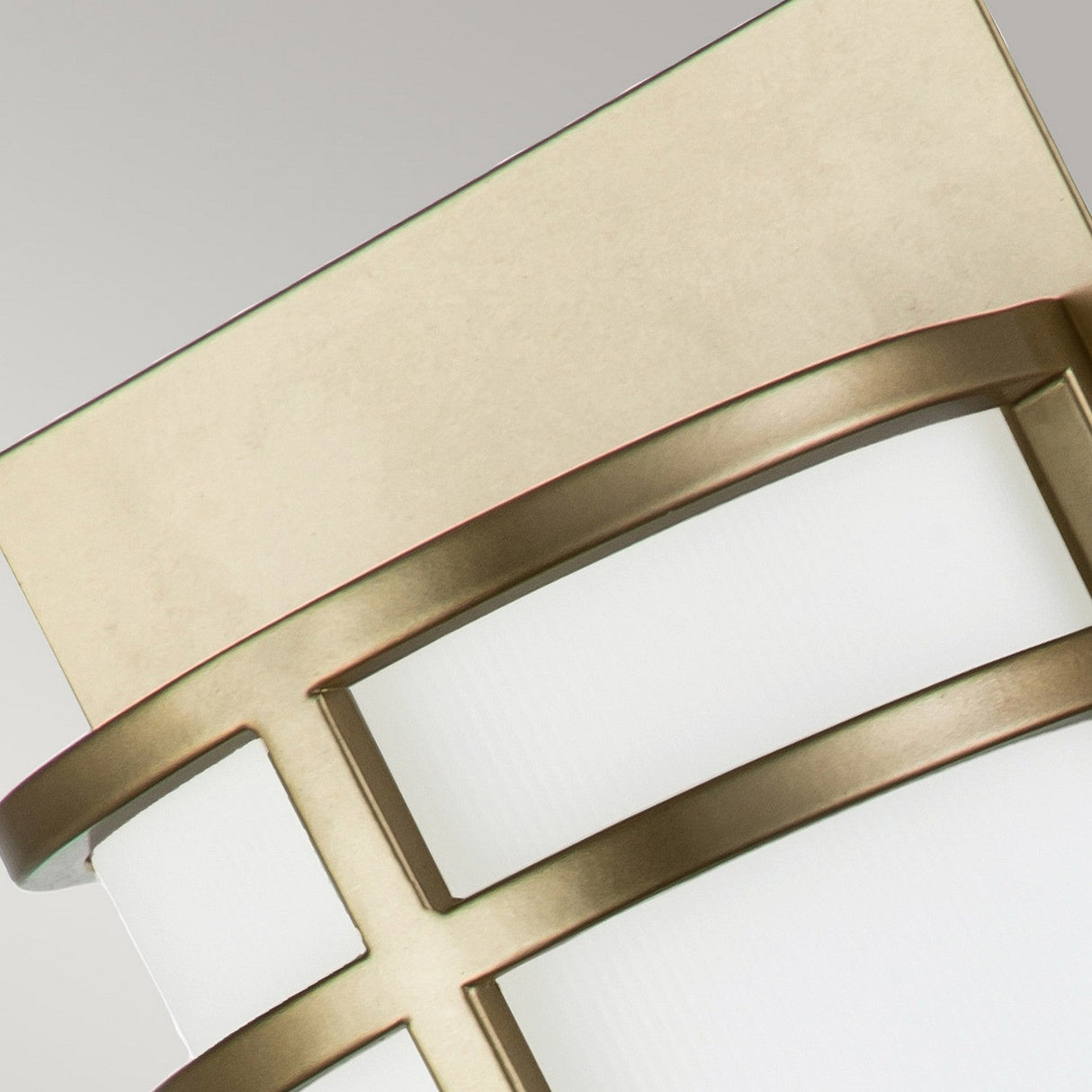 A close-up view of the Fusion 2 Wall Light - Painted Natural Brass showcases its modern geometric design. It features smooth, curved lines and a rectangular backplate, all supporting a white cylindrical shade crafted from opal glass, set against a plain background.