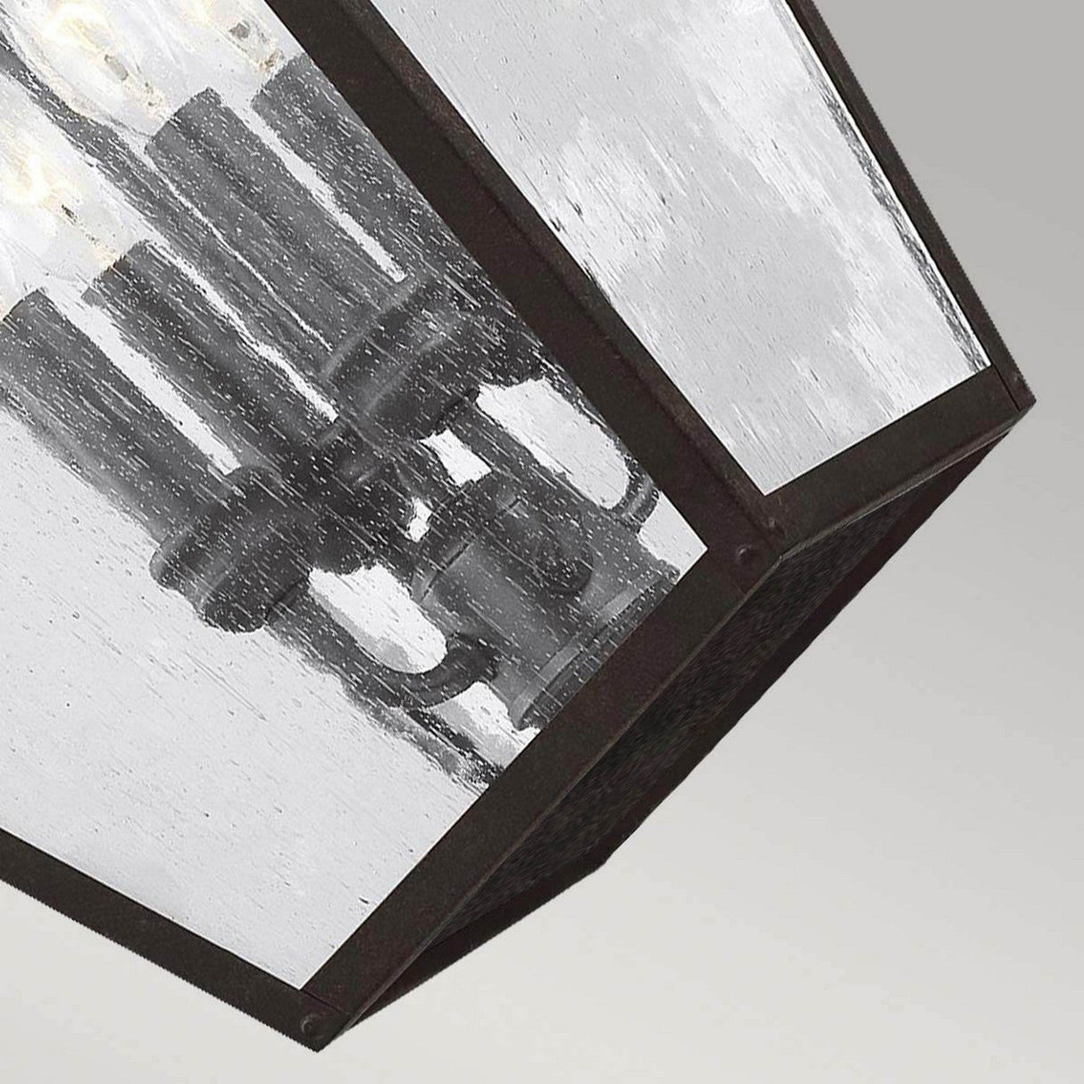 Close-up of the Galena 4 Light Outdoor Chain Lantern, featuring a geometric design with glass panes and a metal frame. Visible bulbs shine through the seeded glass, exuding a modern industrial vibe. Perfectly suited for any setting, its backdrop is a plain, muted gray.