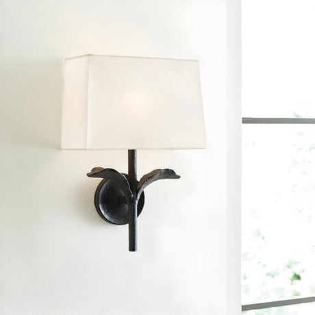 The Georgia 1 Light Wall Light, in its aged iron finish with a white cotton shade, is a wall-mounted fixture showcasing a floral-like design. It stands out beautifully against a light-colored wall near a window, where soft natural light highlights its handcrafted elegance.