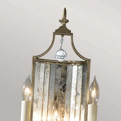 The Gianna 3 Light Wall Light in Gilded Silver features sophisticated chandelier elements with a mirrored backplate and antique detailing. Its intricate gold frame, paired with three candle-style bulbs, beautifully highlights the hand-cut crystals. A crystal pendant at the top enhances the light reflection, creating a luxurious atmosphere.