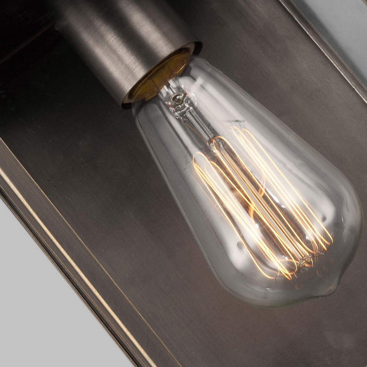A close-up view of an illuminated Glenview 1 Light Outdoor Small Wall Lantern in antique bronze reveals the vintage-style Edison bulb with visible filaments, set against a gray background. This light emits a warm glow that highlights the intricate design of the bulb's internal structure.