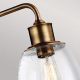 Close-up of the Hobson 3 Light Island Light in aged brass, showcasing its antique style with seeded glass shades. The spotted glass resembles water droplets, revealing an Edison bulb inside that emits a warm glow. The background is a soft gray.