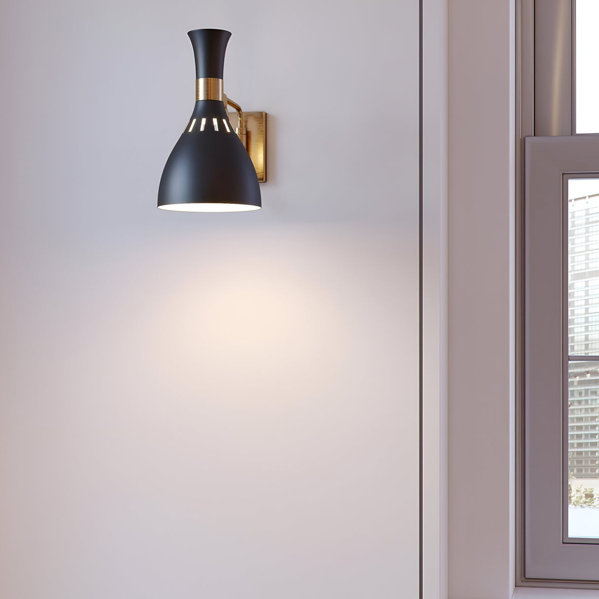 The Joan 1 Light Wall Light in Midnight Black with a Burnished Brass bracket casts light on the pale gray wall. To the right, a window reveals a city skyline under a clear sky.