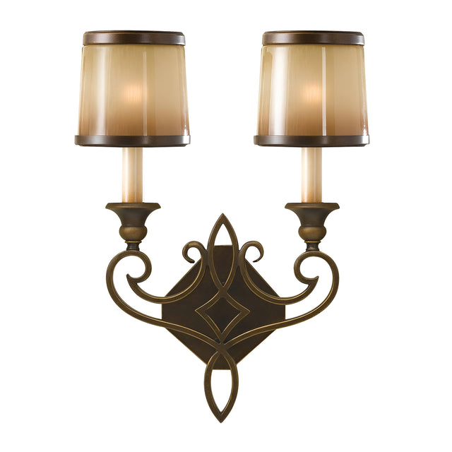 The Justine 2 Light Wall Light in Astral Bronze features two candle-shaped light fixtures, each adorned with a beige cylindrical lampshade. Its ornate dark metal design highlights traditional craftsmanship with a central diamond shape and elegant curved elements.