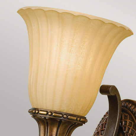 A close-up image of a Kelham Hall 1 Light Wall Light - Firenze Gold/British Bronze reveals a textured, fluted amber glass shade. The fixture features intricate detailing in a Firenze Gold finish, providing an elegant and classic look.