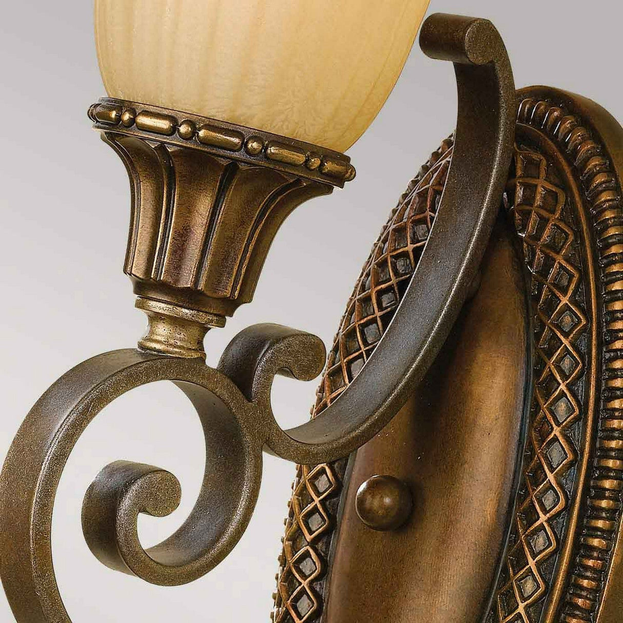 Close-up of the Kelham Hall 1 Light Wall Light with an ornate British Bronze finish. The lamp showcases a detailed scroll design paired with an amber glass shade, all mounted on an oval backplate featuring textured detailing.