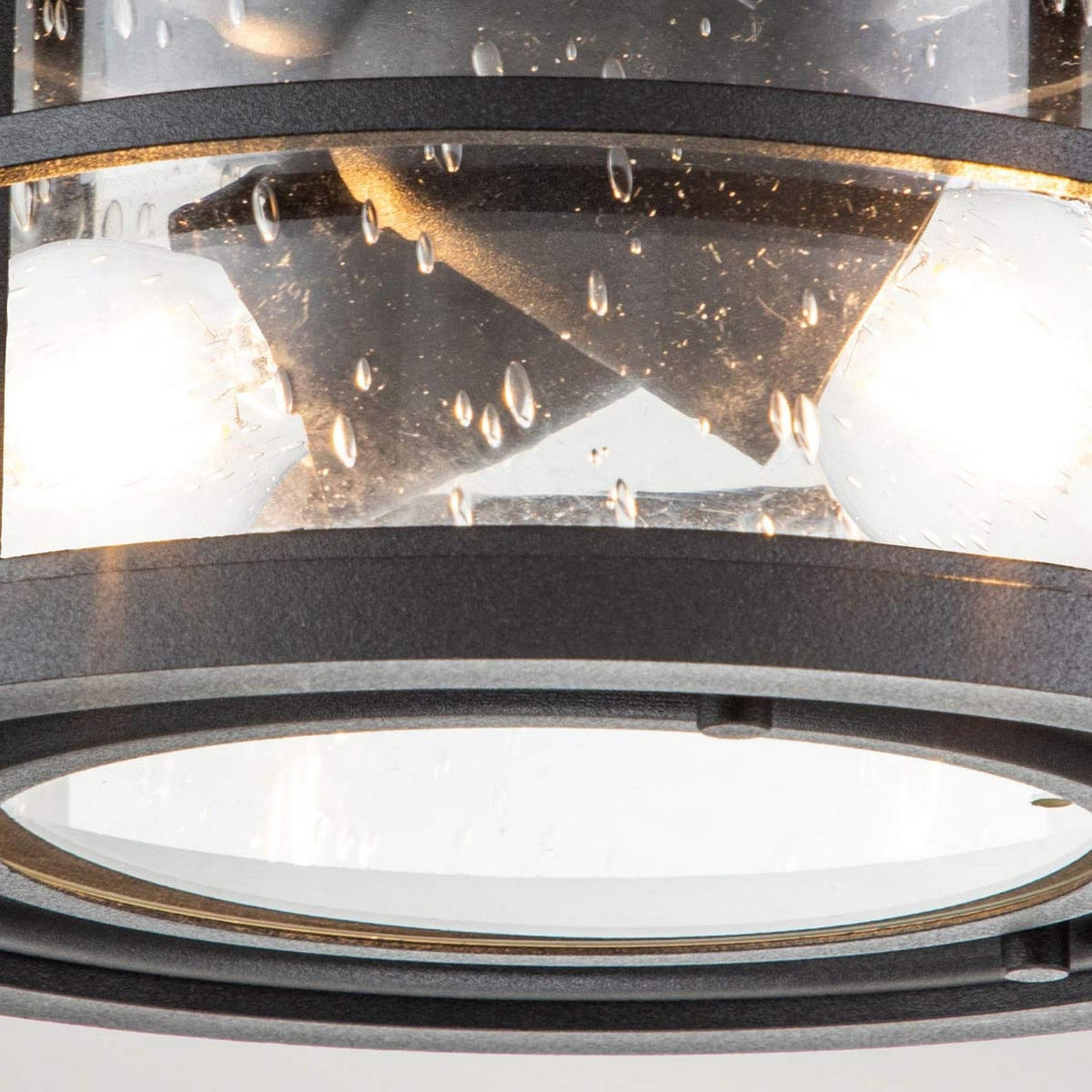 A detailed view of the Lighthouse 2 Light Outdoor Flush - Textured Black reveals a solid brass cage surrounding the fixture. The glass, with its raindrop texture, perfectly accentuates the textured black finish while housing two bulbs that emit a warm glow, adding an aesthetic touch to its design.