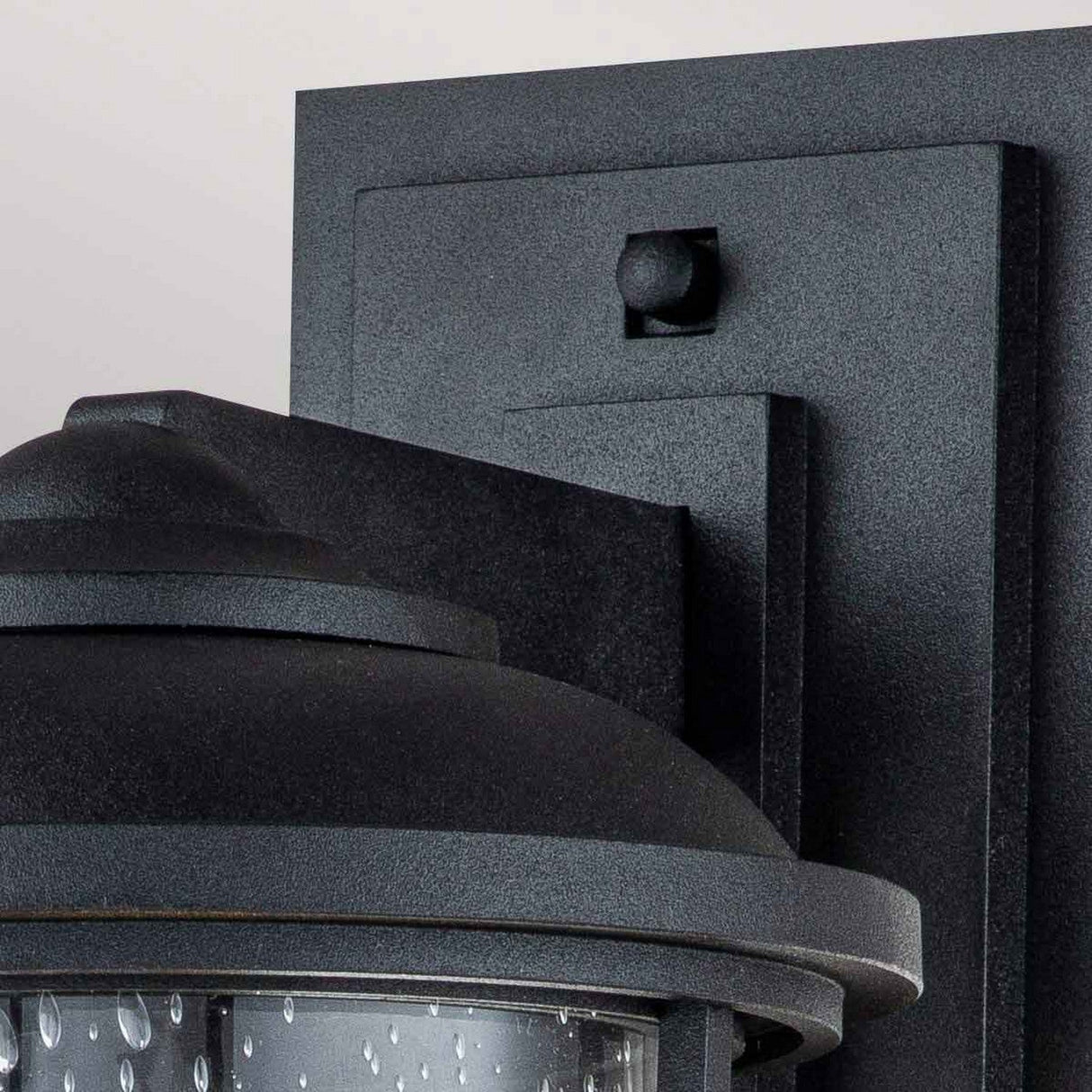 Close-up of the Lighthouse 1 Light Outdoor Medium Wall Lantern in textured black. This fixture showcases a simple, modern design with a clear glass enclosure featuring water-droplet texture, mounted on a flat rectangular backplate that subtly integrates nautical design elements.