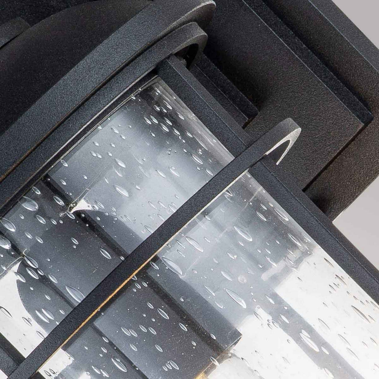 Close-up of the Lighthouse 1 Light Outdoor Medium Wall Lantern showcasing its textured black frame. The glass cylinder, sprinkled with water droplets, suggests a recent rain, set against a soft gray background.