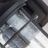 Close-up of the Lighthouse 1 Light Outdoor Medium Wall Lantern showcasing its textured black frame. The glass cylinder, sprinkled with water droplets, suggests a recent rain, set against a soft gray background.