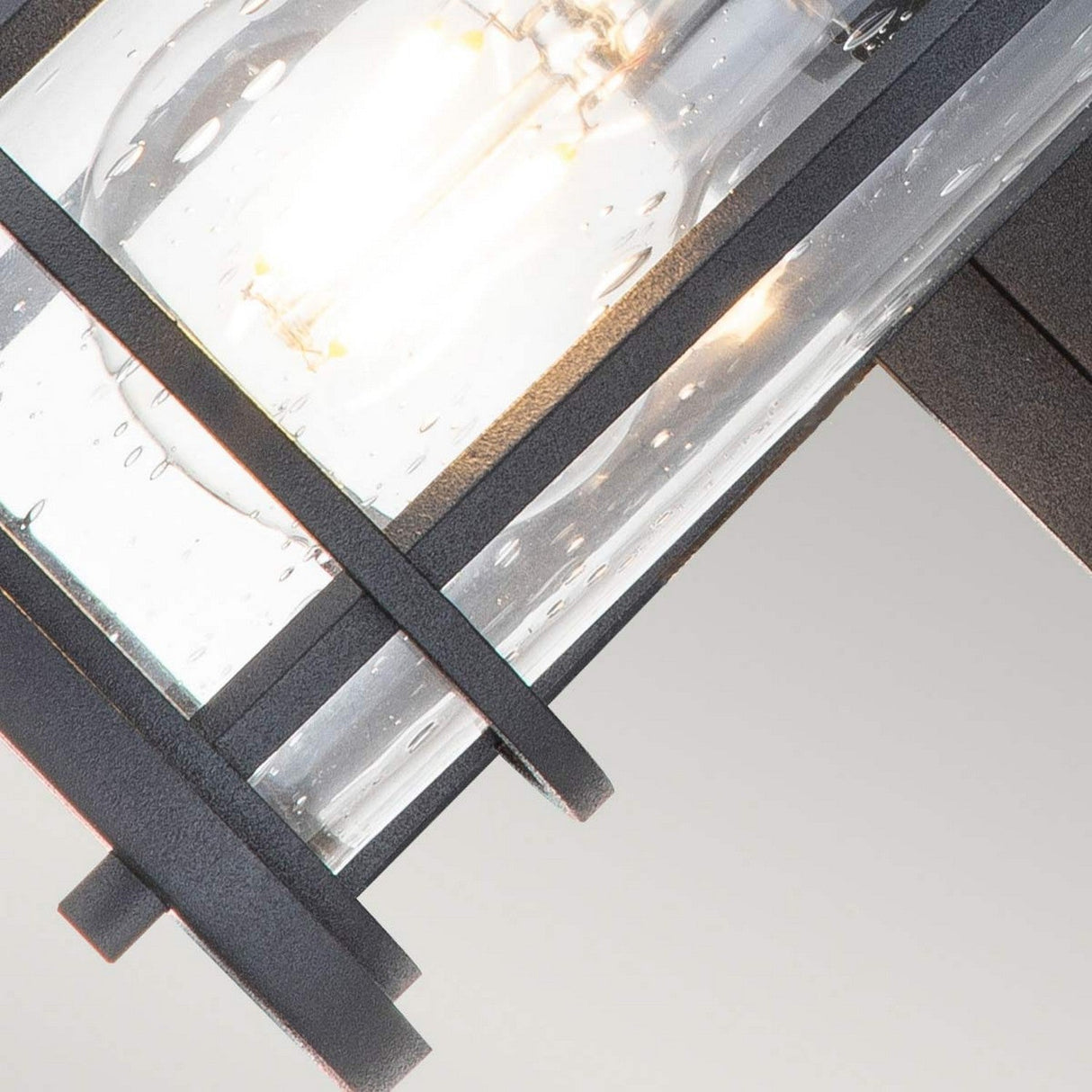 A close-up of the Lighthouse 1 Light Outdoor Small Wall Lantern in textured black metal, featuring clear glass panels. The illuminated bulb inside casts a warm glow that beautifully reflects and dances off the glass.