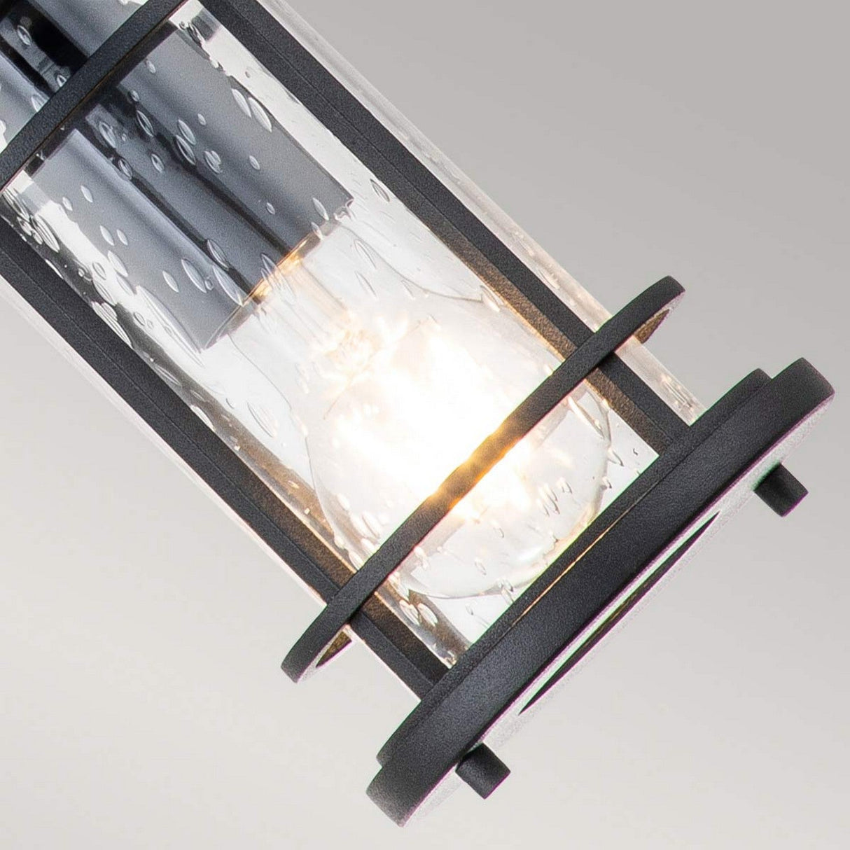 The Lighthouse 1 Light Outdoor Small Chain Lantern in Textured Black features a lit bulb inside a cylindrical fixture covered with clear seeded glass, giving it a bubbled look. This is enhanced by the frame's matte black metal finish, while the soft gray gradient background adds to its elegant nautical lantern appearance.