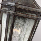 Close-up of the Londontowne 1 Light Outdoor Wall Lantern in weathered chestnut: a vintage-style piece featuring a textured seeded glass enclosure. The metal frame shows signs of rust and patina, while a warm light softly glows inside, casting gentle illumination.