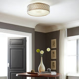 A modern room with a round wooden table holding a white vase and flowers. The wall features framed sketches, and the space is illuminated by the Lucia 6 Light Flush in Burnished Silver from Luxury Lighting Solutions. A black door and white curtains complete the backdrop.