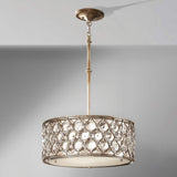 The Lucia 3 Light Chandelier - Burnished Silver is a decorative ceiling fixture with a drum-shaped shade, featuring a metallic frame in burnished silver and adorned with large, clear Bauhinia crystal accents. It elegantly hangs from a single rod against a plain, light gray backdrop.