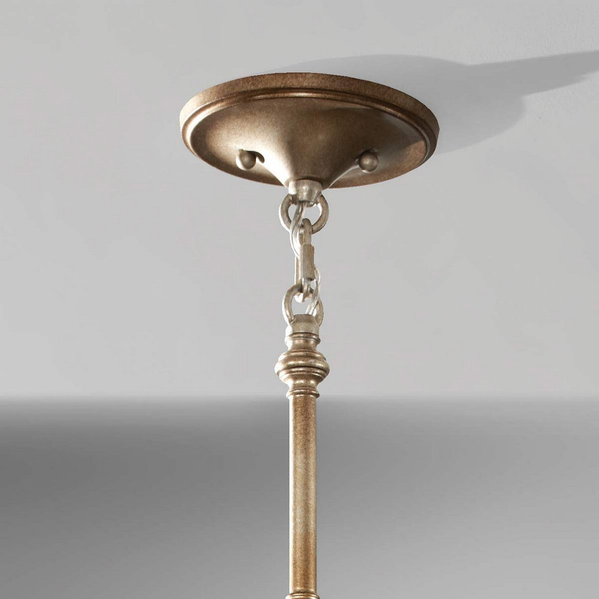 Close-up of a ceiling-mounted metallic light fixture, showcasing a circular base connected by a chain. The fixture resembles the Lucia 3 Light Chandelier - Burnished Silver and hangs from the ceiling against a light gray background.
