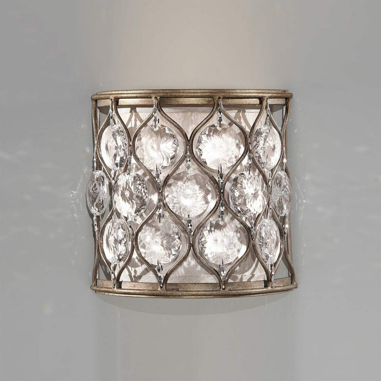 The Lucia 1 Light Wall Light in burnished silver features a decorative cylindrical metal frame adorned with interconnected, leaf-shaped Bauhinia crystals. These crystals beautifully catch and diffuse light, casting a sparkling effect against the neutral backdrop.