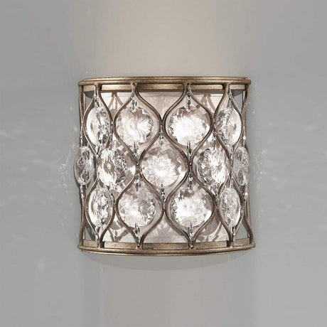 The Lucia 1 Light Wall Light in burnished silver features a decorative cylindrical metal frame adorned with interconnected, leaf-shaped Bauhinia crystals. These crystals beautifully catch and diffuse light, casting a sparkling effect against the neutral backdrop.