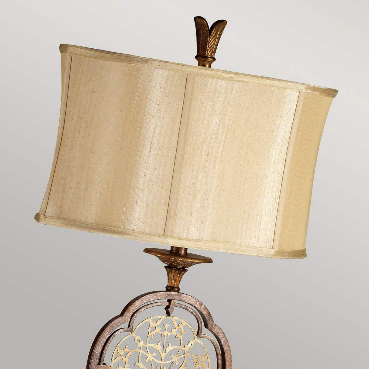 The Marcella 1 Light Floor Lamp - Bronze exudes vintage glamour with its cream-colored, slightly curved lampshade. Its base showcases ornate metalwork adorned with filigree details and floral patterns, topped with a leafy finial.
