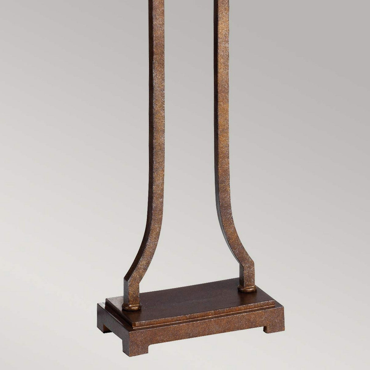 Close-up of the Marcella 1 Light Floor Lamp in bronze, showcasing its metal stand with a square base and two vertical, curved supports. The rustic, weathered finish on the metal adds vintage glamour against a plain, light gray background.