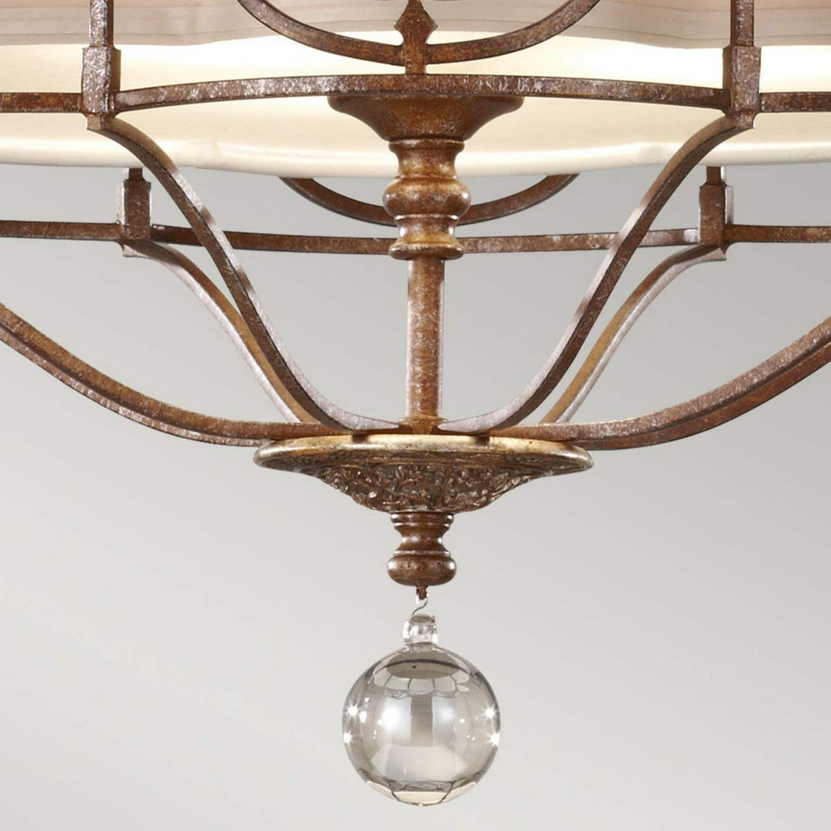 Close-up of the Marcella 3 Light Chandelier in bronze, featuring an intricate filigree design and a spherical glass pendant light that offers ambient illumination. Its curved arms and soft beige shade stand elegantly against a plain light gray background.