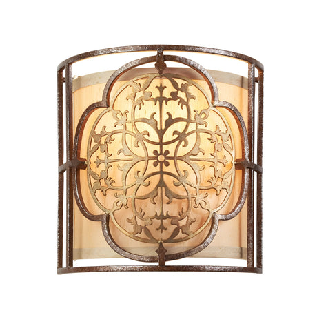 Introducing the Marcella 1 Light Wall Light in bronze, featuring an ornate, symmetrical metal design on its front. The warm light gently radiates through a glass or frosted backdrop, showcasing an intricate filigree pattern that creates an elegant and vintage ambiance.