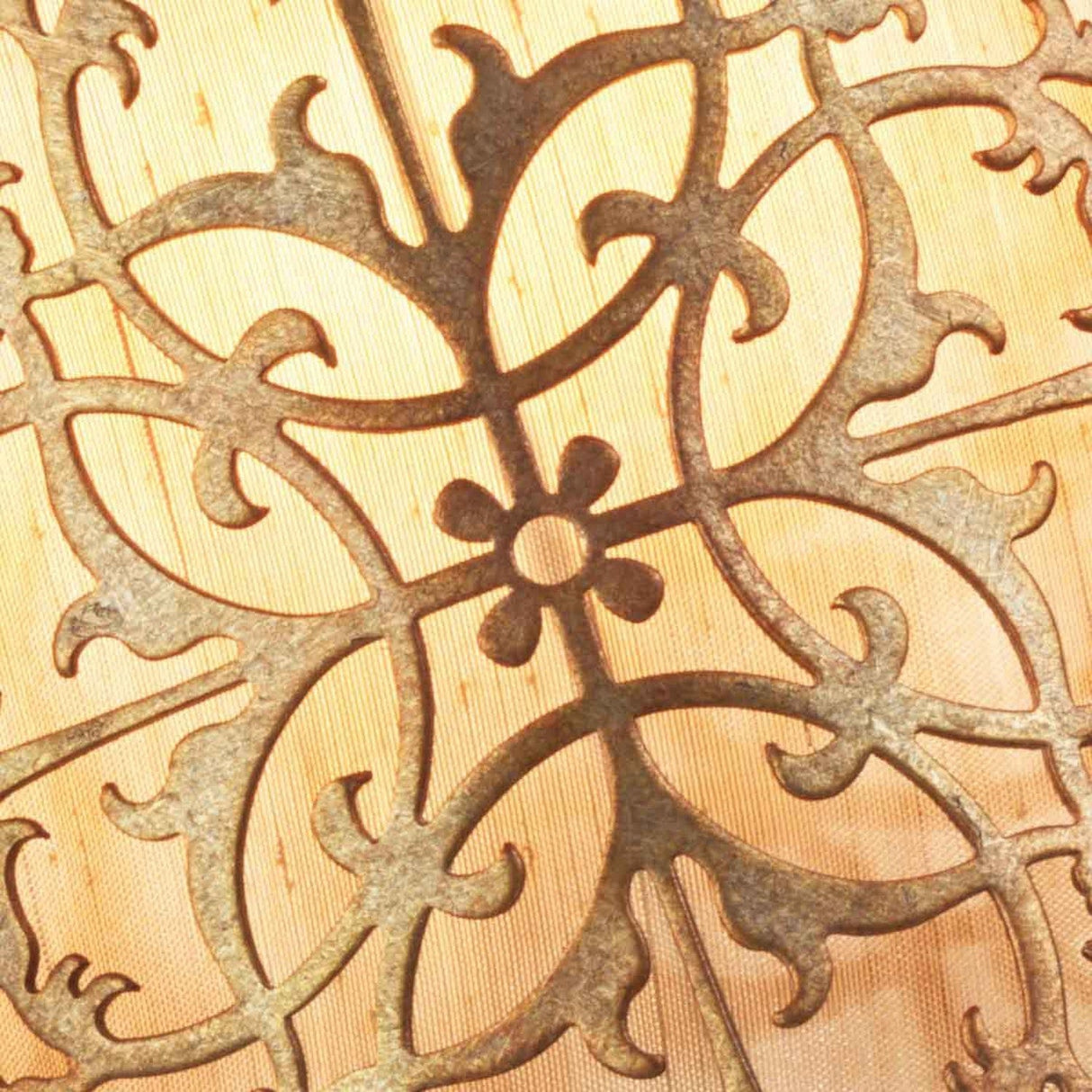 Close-up of the Marcella 1 Light Wall Light in bronze, showcasing its ornate grille with a filigree design that casts intricate shadows on the wooden surface behind it. The pattern includes symmetrical curves and a central floral motif.