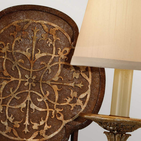 A close-up of an ornate, vintage-style metal wall decoration with intricate filigree detailing features floral patterns. The view is partially obscured by the Marcella 2 Light Wall Light - Bronze, which boasts a beige lampshade and sits atop a lamp with an aged golden finish. The scene evokes a classic, luxury home decor aesthetic.