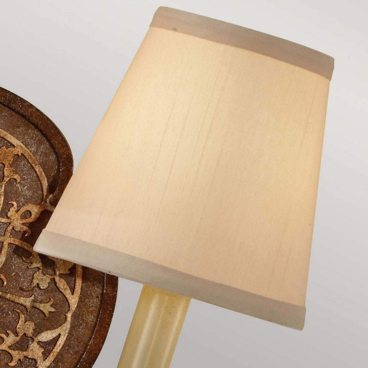 A detailed view of the Marcella 2 Light Wall Light in bronze, showcasing its elegant design with a beige fabric lampshade. It's positioned beside a decorative wooden panel adorned with intricate floral filigree carvings against a neutral gray background.