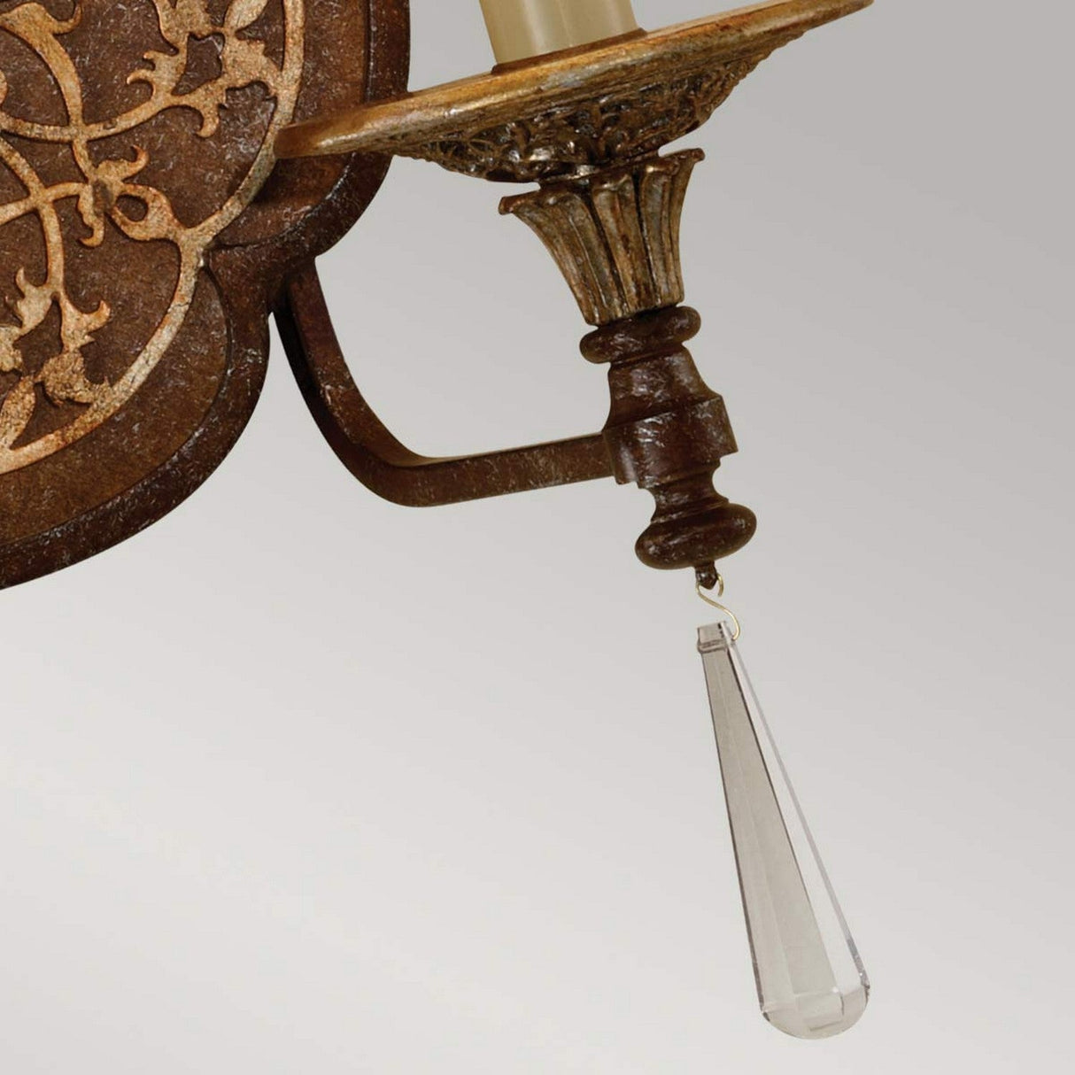 The Marcella 2 Light Wall Light - Bronze showcases a close-up of its ornate design, featuring intricate metalwork with filigree detailing and two candle-style fixtures. A decorative crystal teardrop pendant dangles from the bottom, enhancing the luxurious touch of this home decor piece. The background is a plain, light-colored wall.