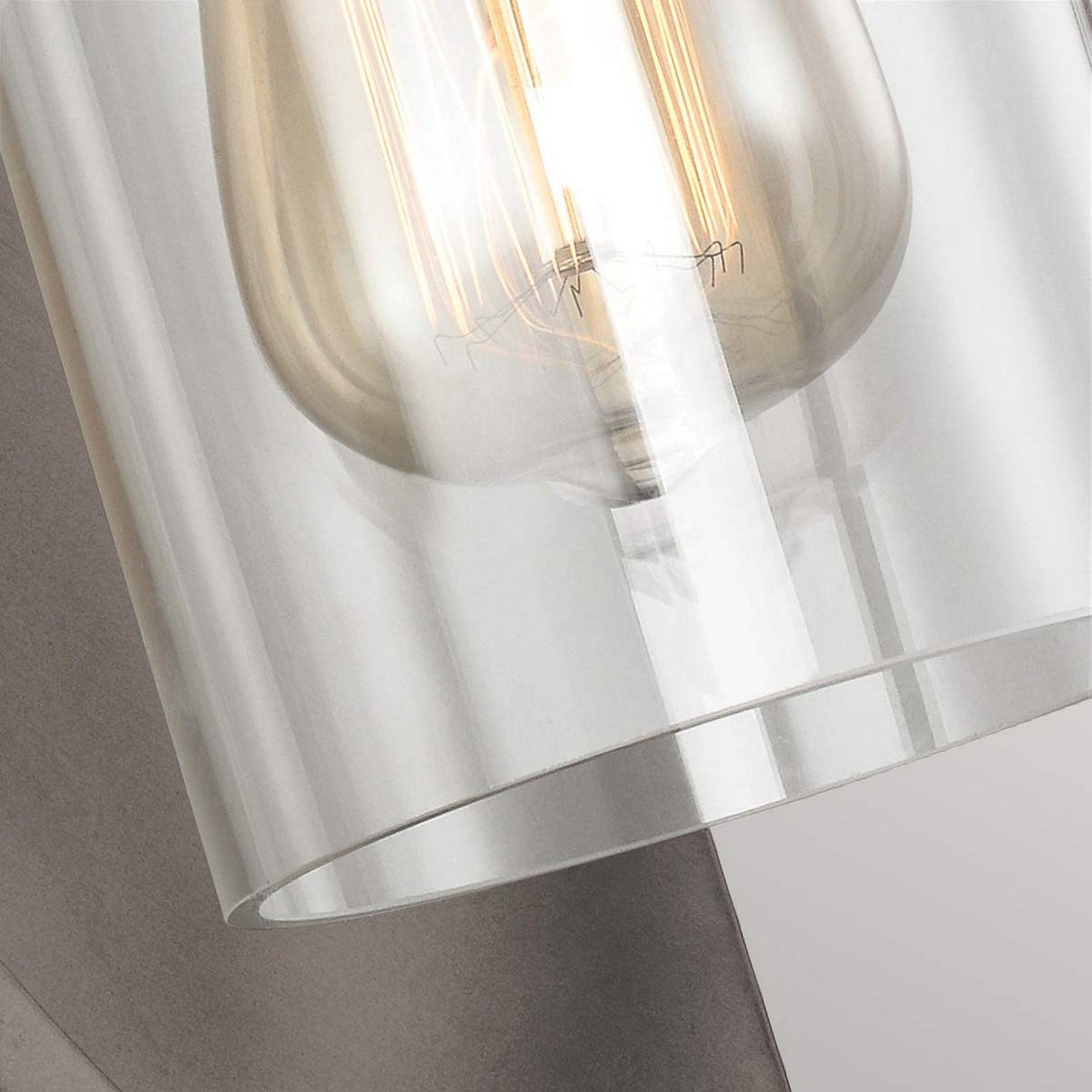 A close-up of the Marlena 1 Light Wall Light in Antique Bronze features a transparent glass light fixture with visible filaments inside the bulb, set against a neutral background that highlights its geometric design.