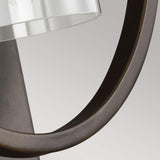 Close-up of the Marlena 1 Light Wall Light - Antique Bronze, showcasing its circular metal frame in an antique bronze finish and a transparent glass shade. Set against a neutral background, it emphasizes its sleek and contemporary geometric design.