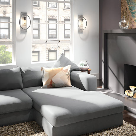 In a contemporary living room, a light gray sectional sofa is complemented by a pillow with a geometric pattern. Sunlight streams through large windows, brightening the ambiance. A fireplace with logs rests beneath Marlena 1 Light Wall Lights in polished chrome, accentuating the vibrant setting.