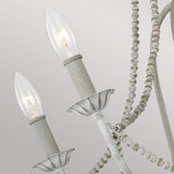 A close-up of the Maryville 6 Light Chandelier, showcasing its rustic elegance with two lit candle-style bulbs. This fixture embodies French Country style with a distressed white finish and decorative beaded chains, set against a soft gray background.