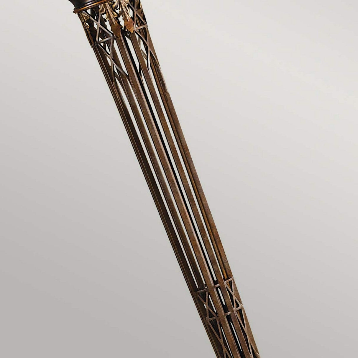 A close-up of the Opera 1 Light Torchiere in Firenze Gold, featuring a tall, slender design with intersecting beams reminiscent of a tower structure set against a simple gray background.