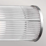 The Paulson 2 Light Bar Light - Chrome, with its cylindrical glass wall sconce and ribbed texture, is installed on a light-colored wall. This design showcases vertical lines and a sleek, modern aesthetic.